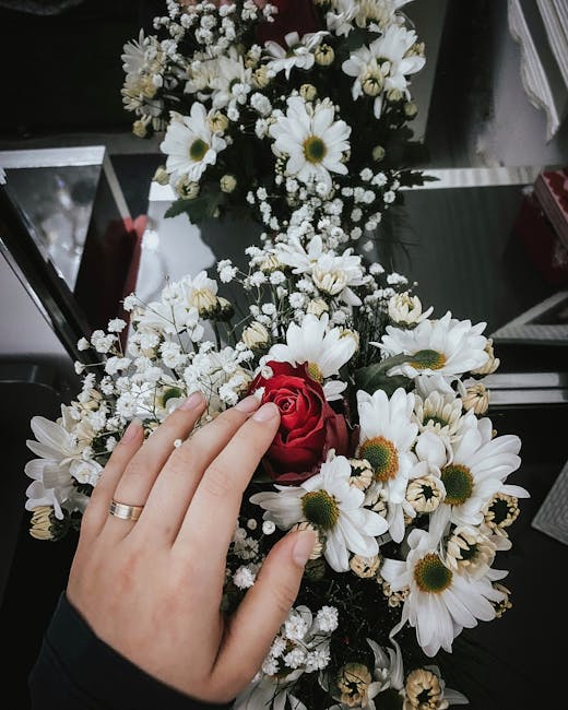 A close-up of a floral arrangement featuring a hand with a silver wedding band gently resting on a bouquet of fresh white daisies with yellow centers, small white baby's breath flowers, and a single r