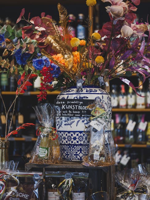 A vibrant floral arrangement featuring pink, purple, and red flowers, including orchids and lilies, interspersed with yellow billy balls and green foliage. The bouquet is displayed in a blue and white