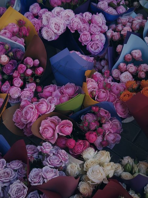 An array of fresh flower bouquets featuring predominantly pink roses with some white and peach roses interspersed. The bouquets are arranged in colorful wrapping paper and placed on a dark surface, wi