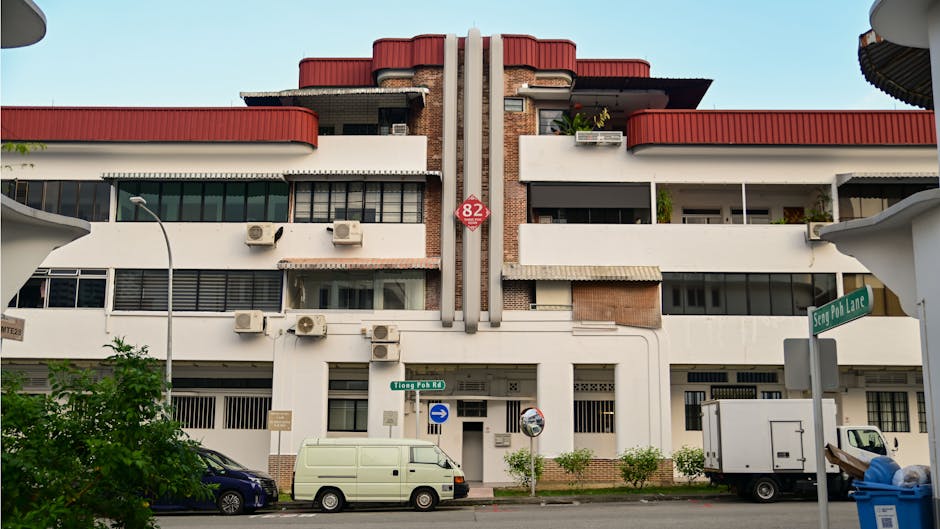 A multi-storey residential building with a modern architectural design featuring white and red-painted sections, large windows, and visible air conditioning units on the exterior. In the foreground, a