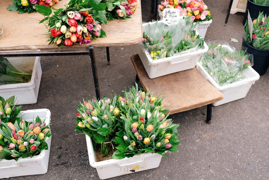 Arrangement of fresh tulip bouquets with pink, white, and orange blooms, placed in white plastic crates and on small wooden tables at an outdoor flower stall. The tulips are in various stages of buddi