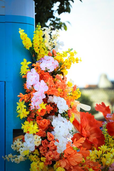 A vibrant floral display featuring a cascading arrangement of fresh flowers attached to a bright blue lamppost. The bouquet includes yellow snapdragons, pink and white orchids, orange roses, and large