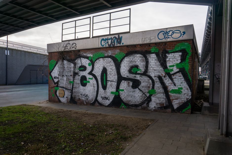A brick wall decorated with colorful graffiti featuring the word 'BOOST' in bold, stylized letters outlined in black, filled with shades of gray and accented with bright green spray paint. The wall is