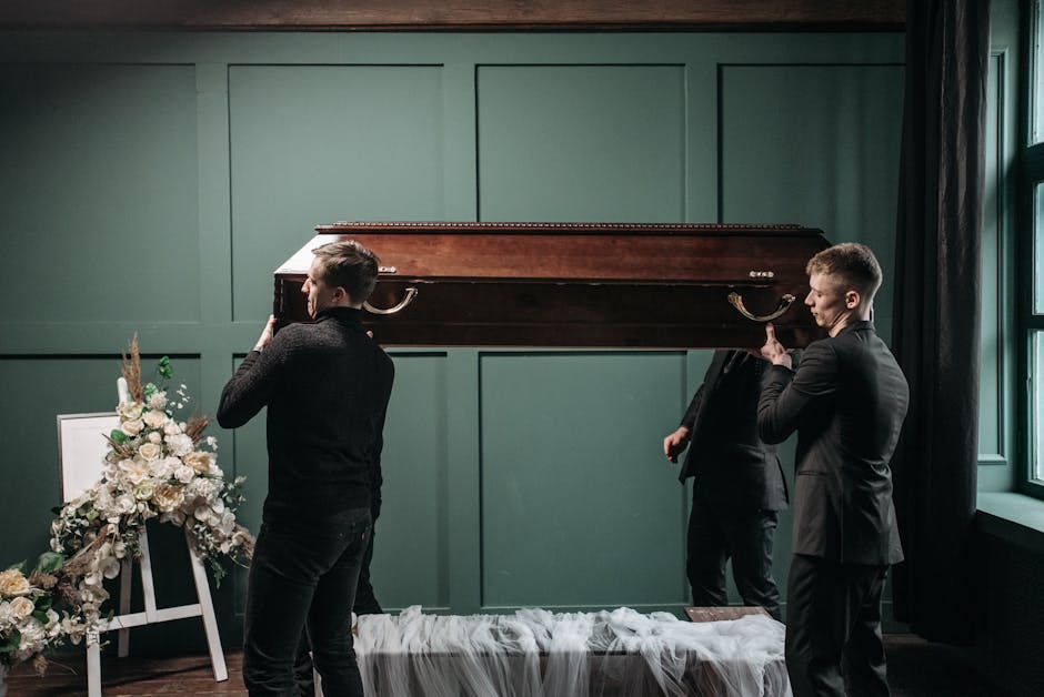 Three men dressed in dark suits are carrying a closed, polished wooden casket with brass handles into a room with dark green paneled walls and a large window. One man is on each side holding the caske