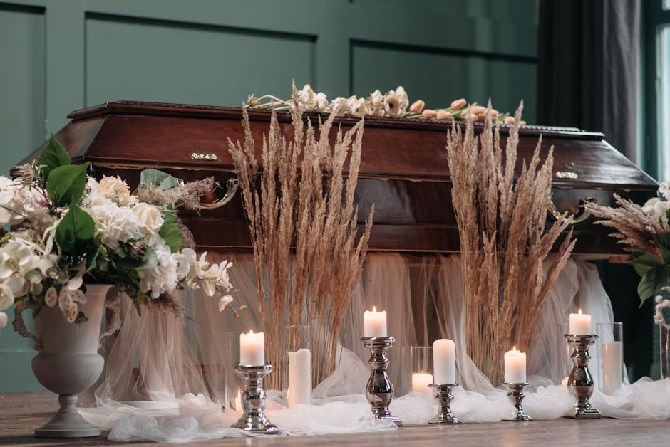 A wooden coffin surrounded by floral arrangements and candles, set on the floor. The flowers include white roses, peonies, and greenery, arranged in white ceramic vases and soft pink wrapping. Tall, d