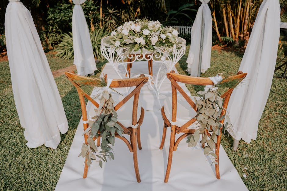 A floral arrangement featuring an abundant bouquet of white roses and hydrangeas, complemented by lush greenery, displayed on a white wrought iron table. The setting is an outdoor garden with sunlight